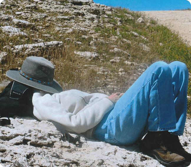 Person relaxing on rocky ground wearing a hat and blue pants.